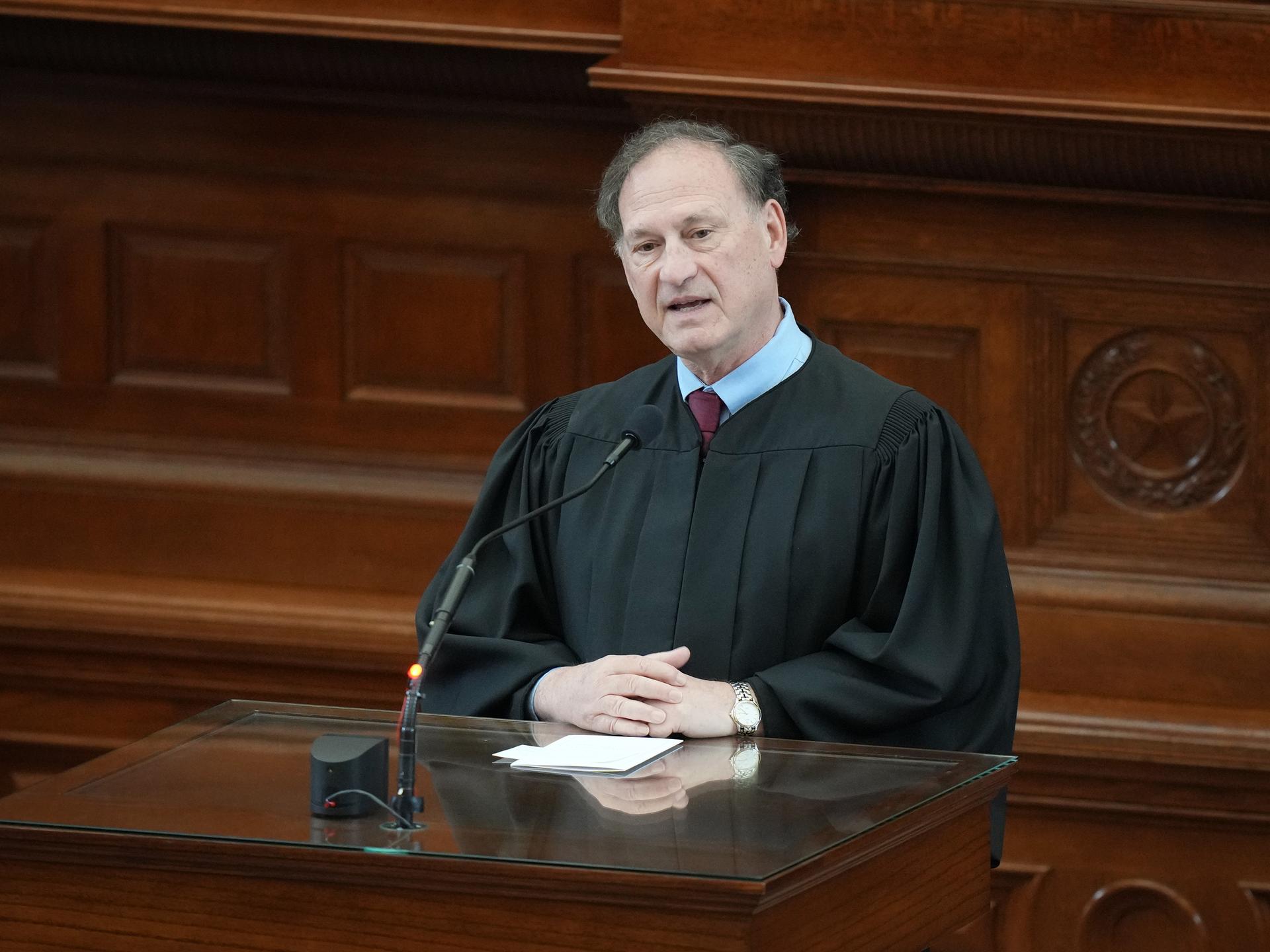 Samuel Alito at microphone, wearing a black judge's robe, with wood paneling behind him.