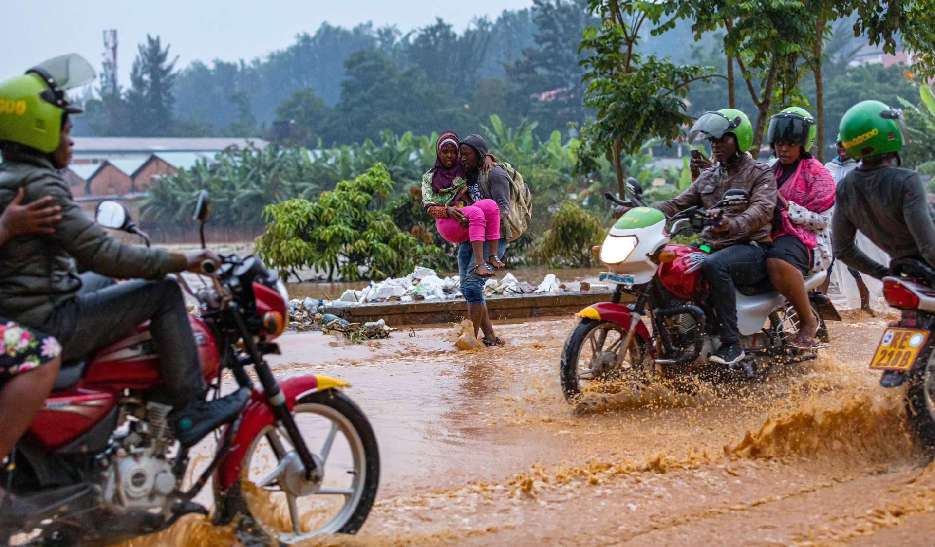 Commuters navigated a muddy, flooded road on foot and motorbike. Behind them, there is a river surrounded by lush green trees.