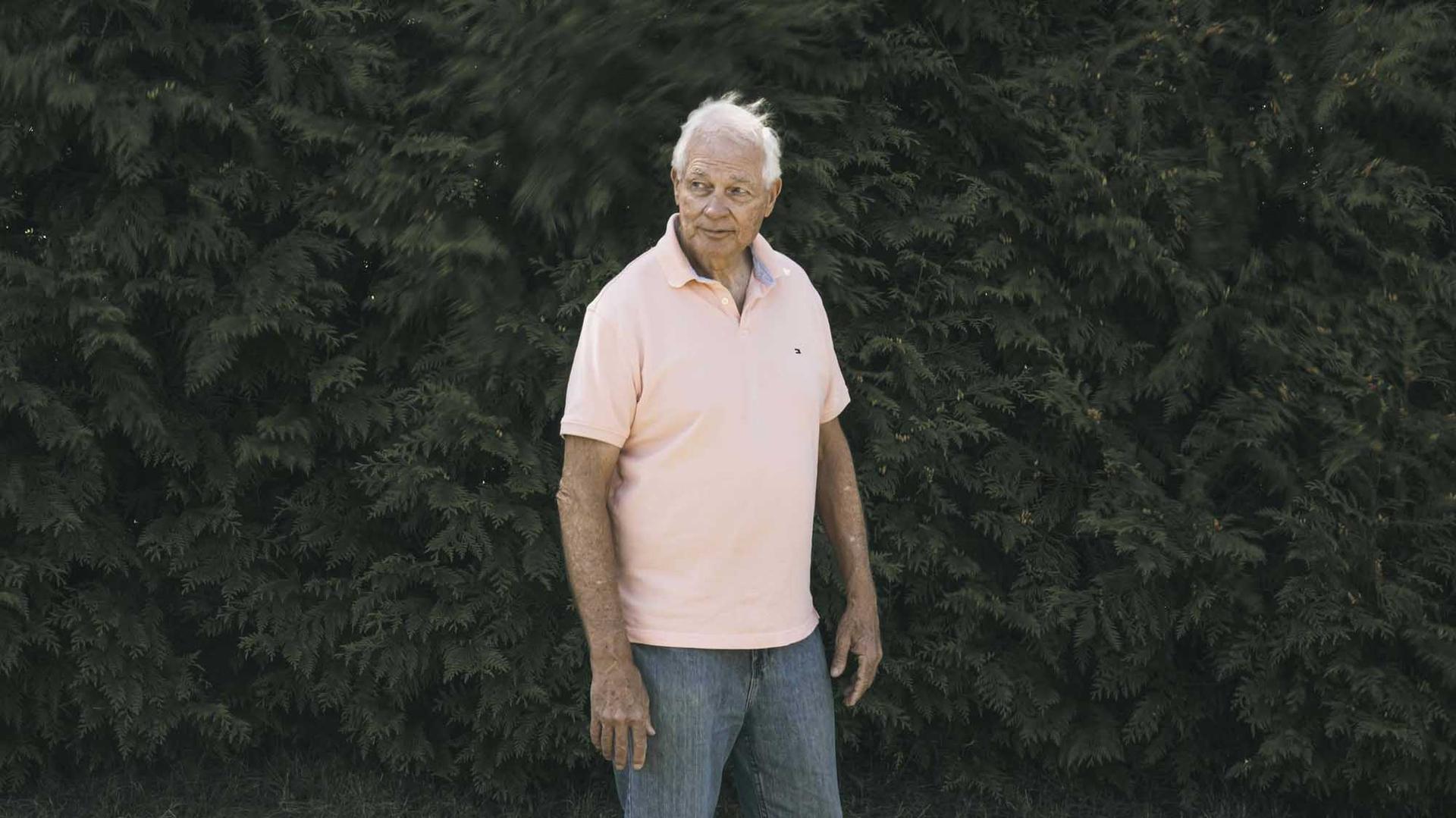 A white, tan man with white hair stands in front of a hedge. He wears a salmon polo.