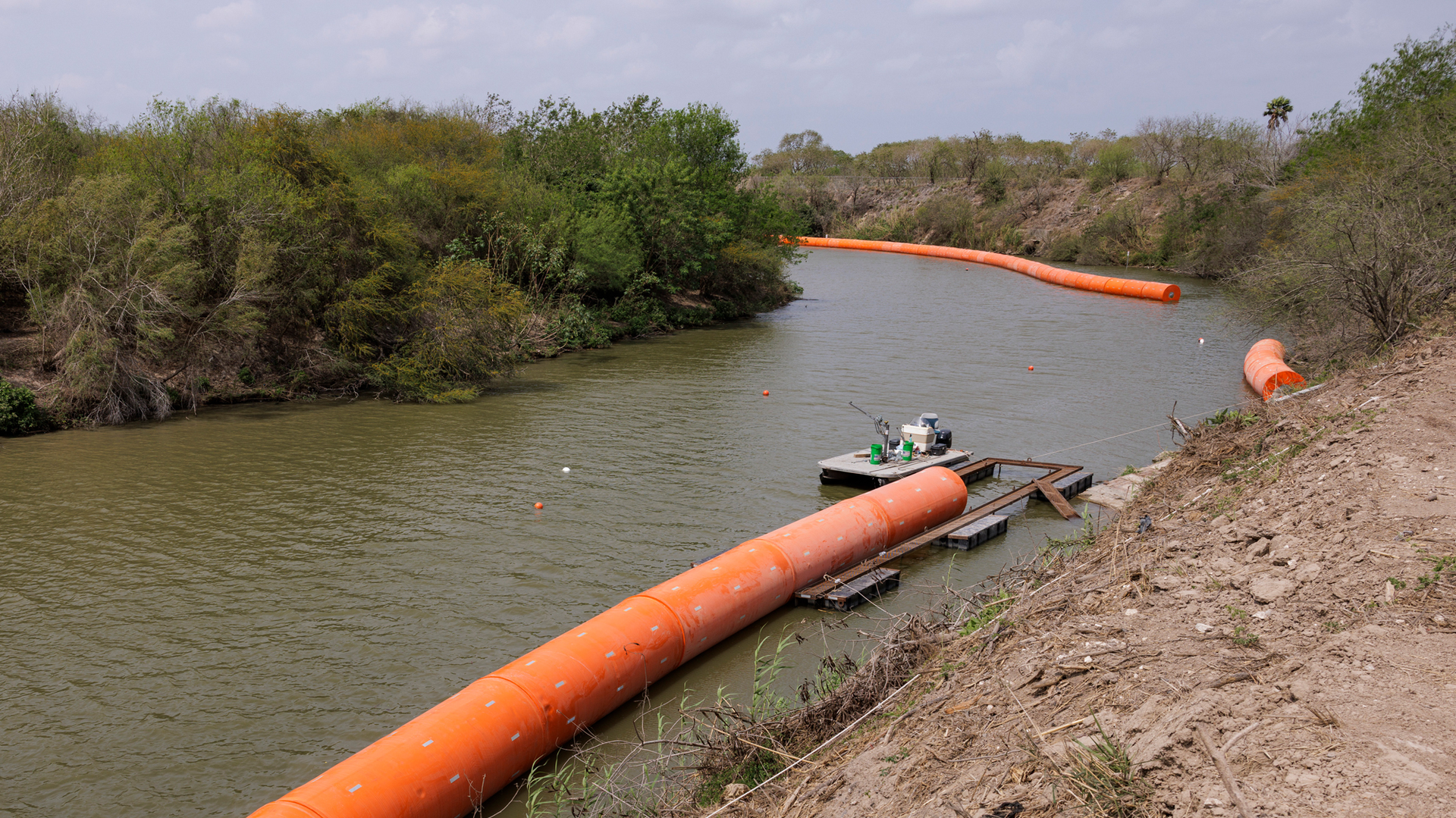 Massive, long, cylindrical orange buoys float on the edge of a river