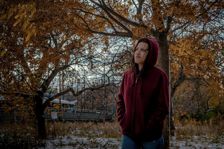 A woman with dark hair in a maroon sweatshirt stares up to the left amid trees with golden leaves.