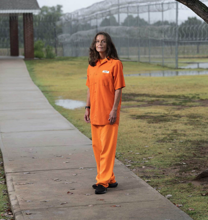 A woman with long brown hair wearing an all-orange outfit stands on a concrete path in a grassy space with a barbed-wire-topped fence in the background.
