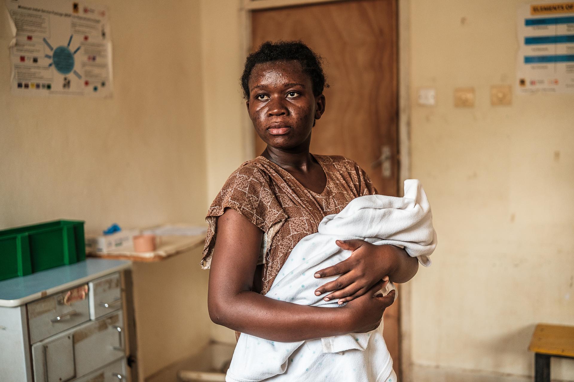 A woman stands holding a blanket-wrapped infant in a hospital room. The room has old, damaged furniture and peeling posters on yellow walls.
