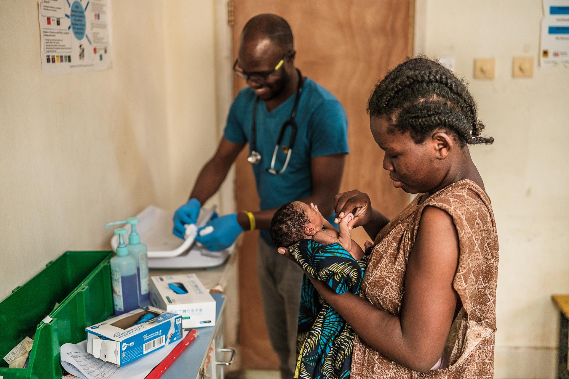 A woman cradles a tiny infant in one hand while holding the baby’s hand with her other hand. They are in a hospital room. In the background, a man with a stethoscope around his neck prepares medical equipment.