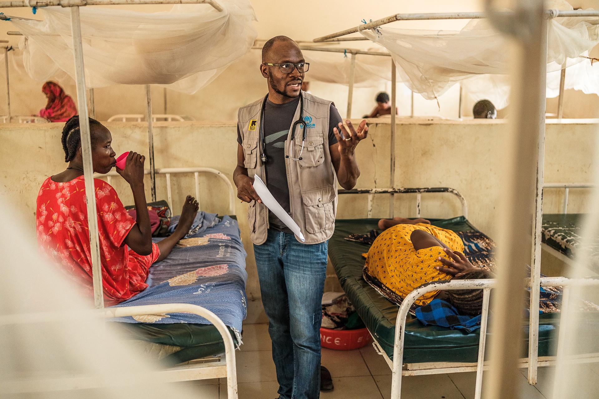 A man with a stethoscope around his neck stands in the center of a hospital room with his hand raised, gesturing to someone outside the frame of the photograph. Around him are numerous hospital beds made up of simple metal frames and thin mattresses, each with a mosquito net hanging overhead. On one bed next to him, a woman sits, drinking from a plastic cup. On the other side of him, a woman sleeps on one of the beds.