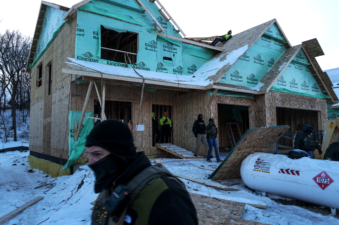 Men in tactical vests and ski masks walk out of the plywood shell of a house. A worker in a high-visibility vest leans against the peak of a roof as others on the ground watch the agents go.