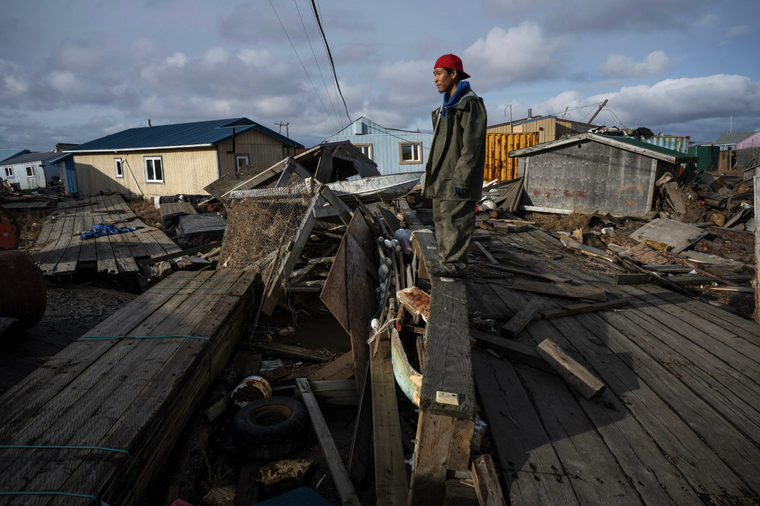 A man stands on a boardwalk railing surrounded by piles of broken wood and debris. Houses sit crookedly behind him and the sky is cloudy.