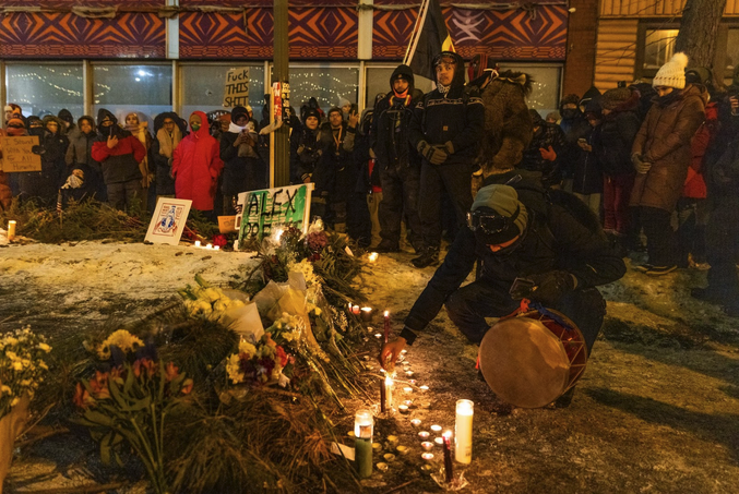 Flowers and candles piled in a semicircle on a ridge of snow. A spray-painted sign says “Alex Pretti,” and a crowd has gathered. One person holds a drum in one hand as they crouch and reach toward a candle with the other hand. 