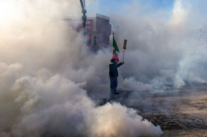 A man holds a Mexican flag and a protest sign in a cloud of tear gas. A street light is just visible behind him, but otherwise the cloud of gas is so thick and tall that the rest of the street is obscured.