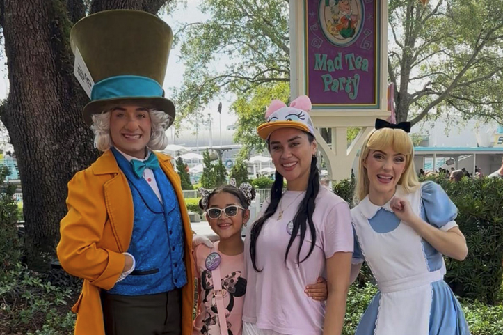 Four people smile and pose for a photo in front of a sign that says “Mad Tea Party”: one person is dressed as the Mad Hatter from “Alice in Wonderland,” one is dressed like Alice, one is a young girl wearing a Mickey Mouse shirt and one is a woman wearing a Daisy Duck visor.
