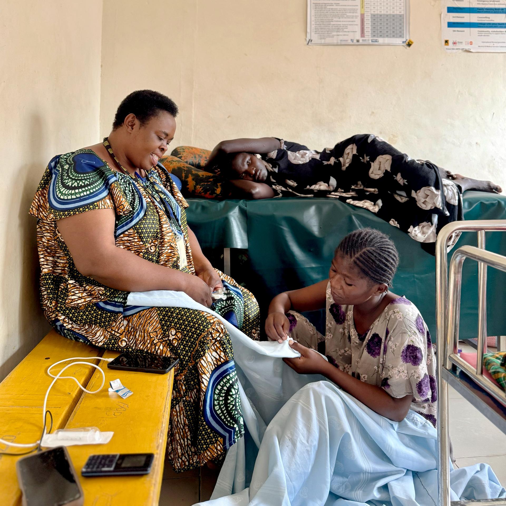 A woman sits on the floor of a hospital room, sewing blue curtains. Another woman sits on a bench in conversation with her. In the background, a third woman lies on a hospital bed.
