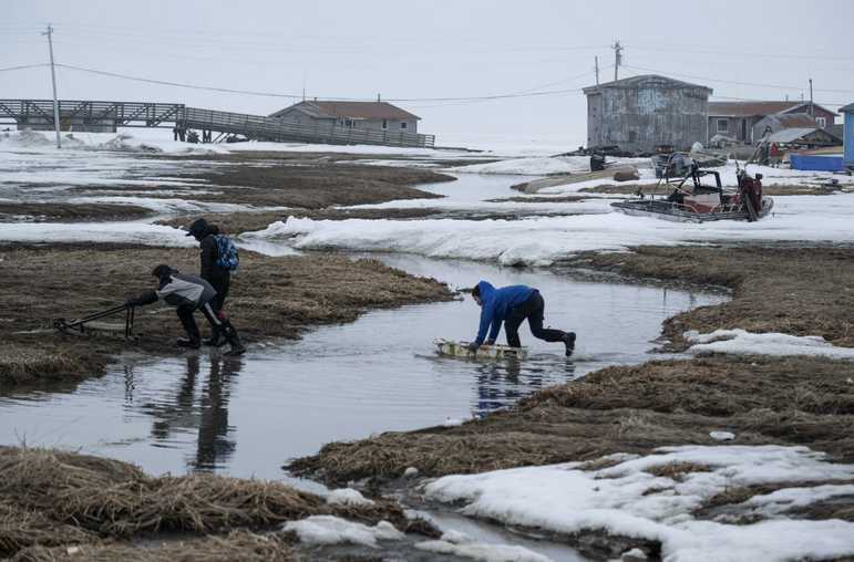 Three kids cross a small river using sleds. A system of boardwalks, small houses, telephone poles and a boat lie in the background. The ground is partially blanketed in melting snow.