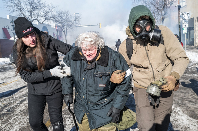 Two people, bundled in winter clothing, support an older woman under her arms. The woman is wincing in pain and holding a cane as the trio moves away from a cloud of tear gas on a snowy urban street.