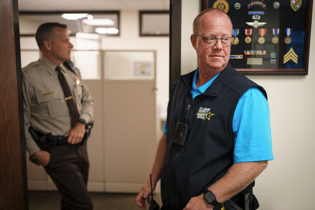 A uniformed sheriff’s officer leans against a doorframe as a sheriff’s officer with a blue shirt under a dark blue vest wearing glasses looks off to the right, inside an office.