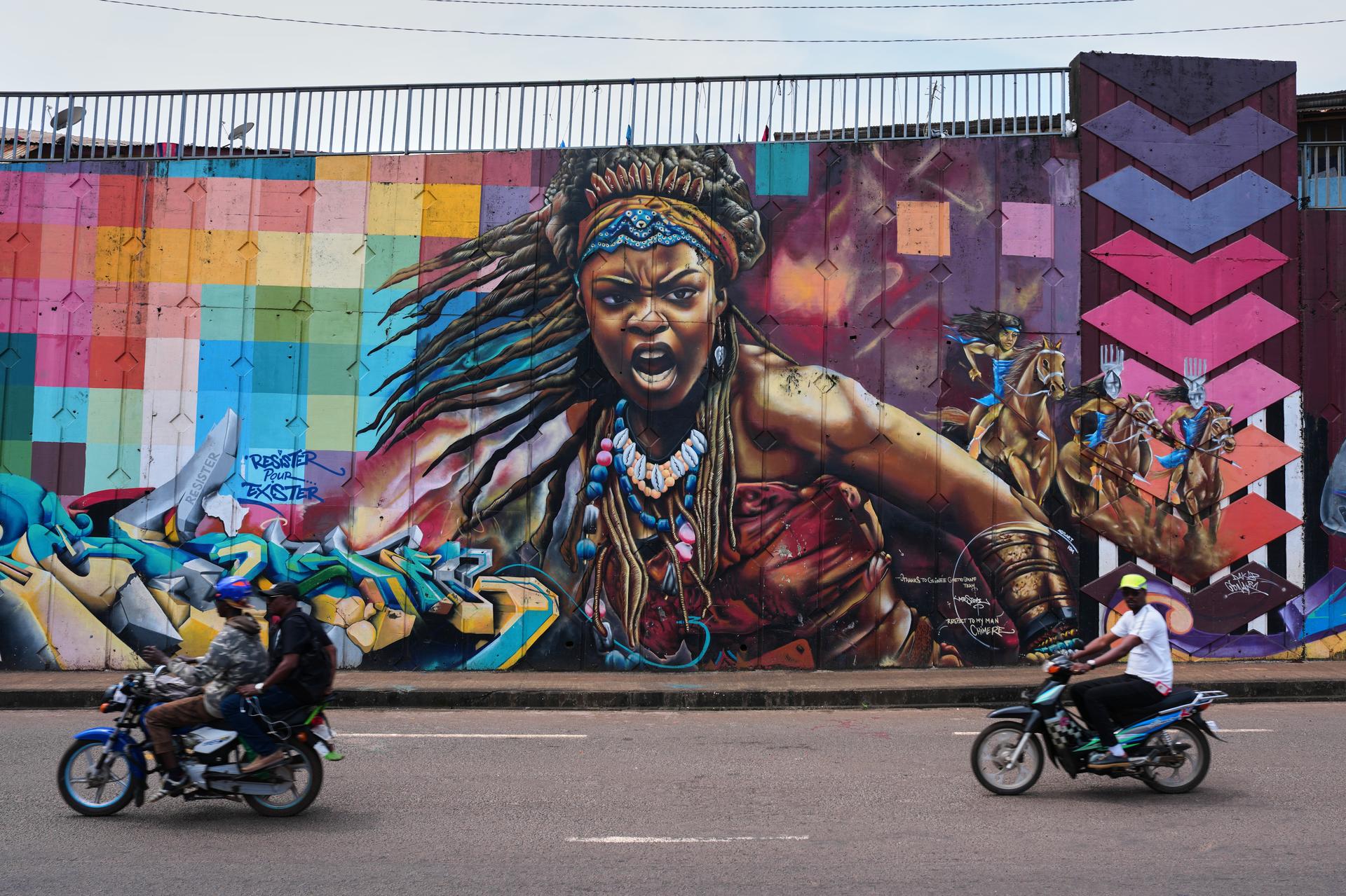 Motorcyclists ride past a mural depicting a powerful woman on a street wall in Conakry, Guinea.