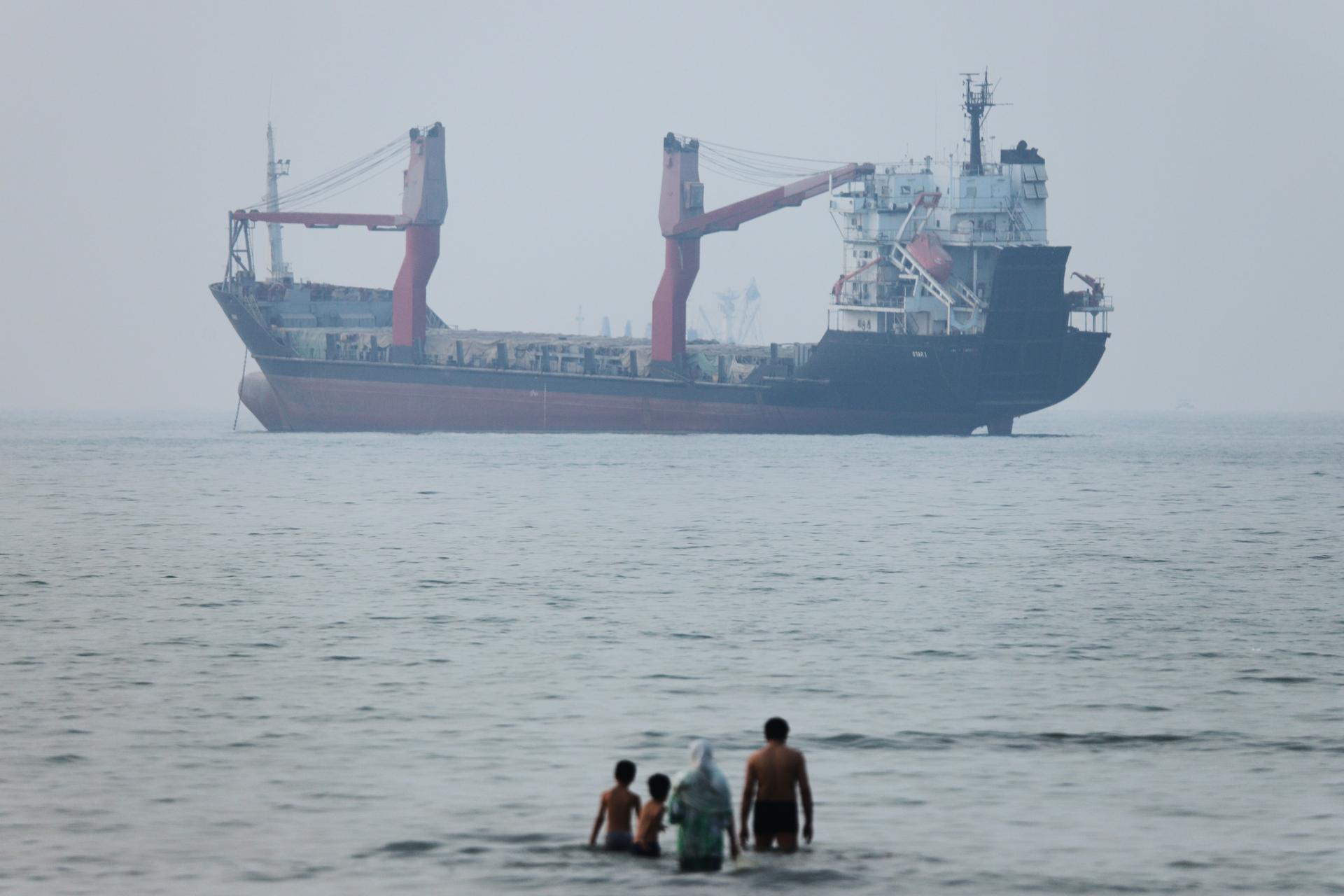 A group of people stands in shallow water as a cargo ship appears anchored in the Strait of Hormuz off Bandar Abbas, Iran, June 30. 