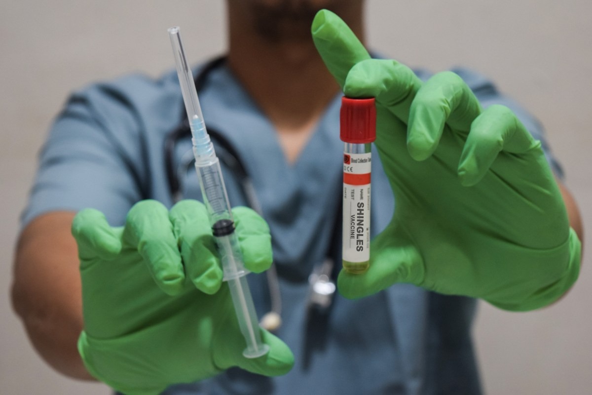 A medical technician wearing green latex gloves holds up a vaccination needle and vial.