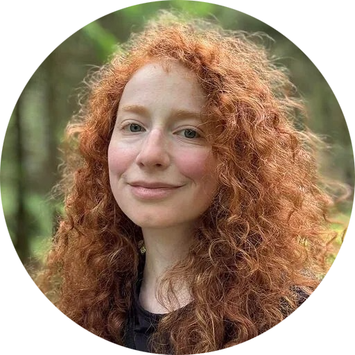 Shayna, a white woman with curly red hair, smiles; out of focus behind her is a green forest.
