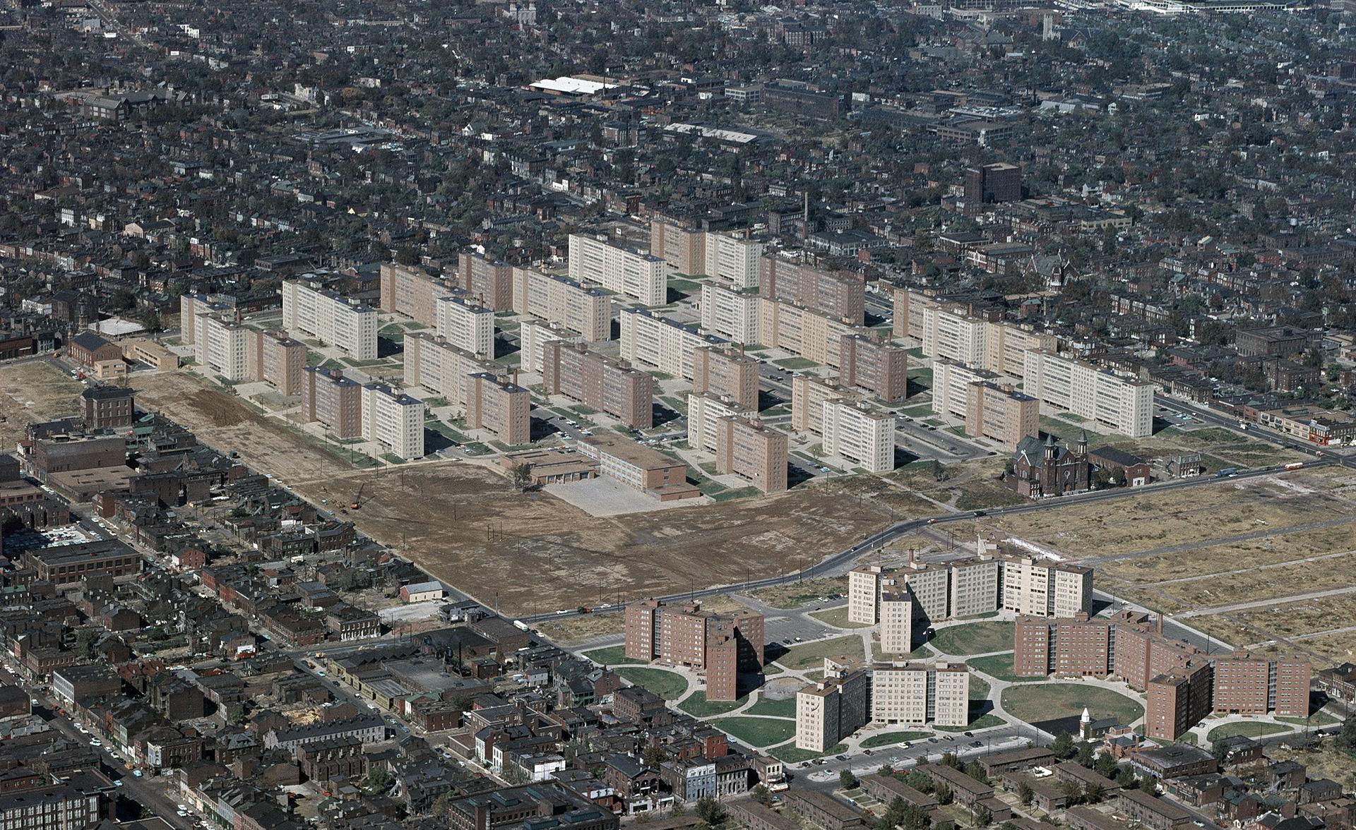 oblique aerial photograph of the Pruitt–Igoe housing complex in St. Louis. Many long, rectangular high-rise apartment blocks are arranged in repeated rows with wide gaps between them, casting dark shadows onto open lawns and paved walkways; surrounding the complex is a tight grid of smaller neighborhood buildings and streets.