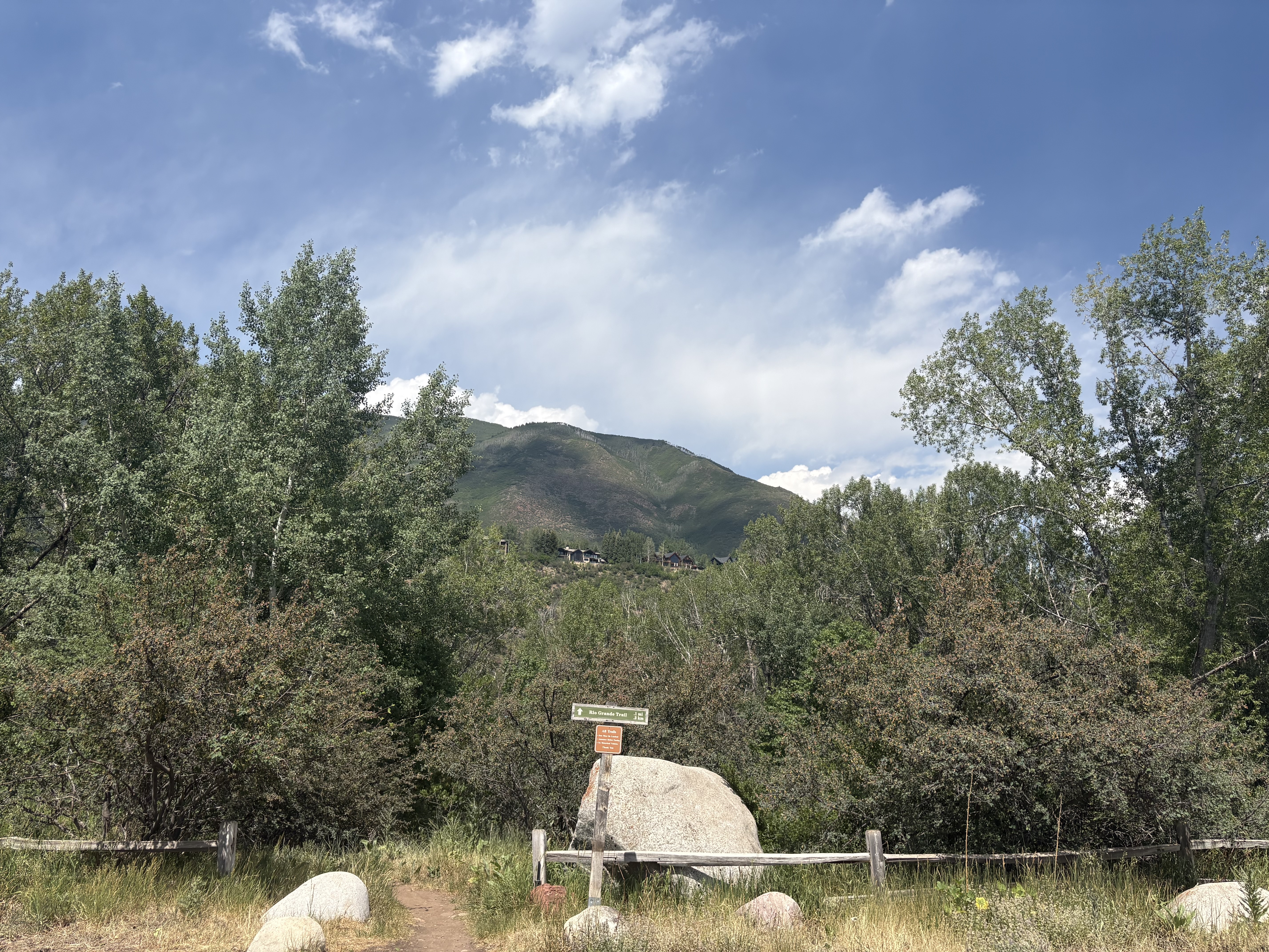 A view of a trailhead beginning behind a fence and disappearing into trees, with tall green mountains in the background.