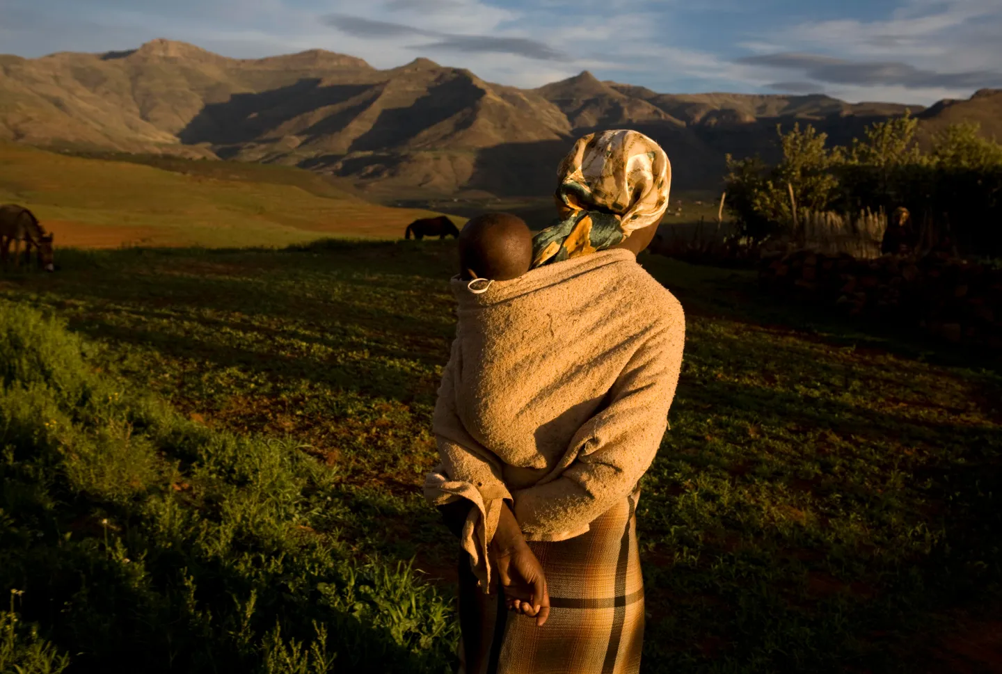 An African mother stands near a plowed field with her baby in a sling on her back.