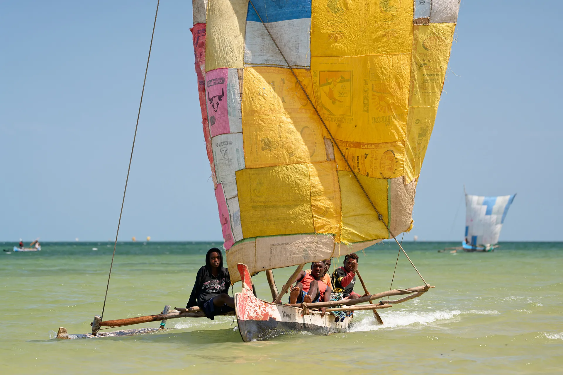 Fishermen carry large, colorful nets inland. Behind them are several colorful boats on the ocean shore.