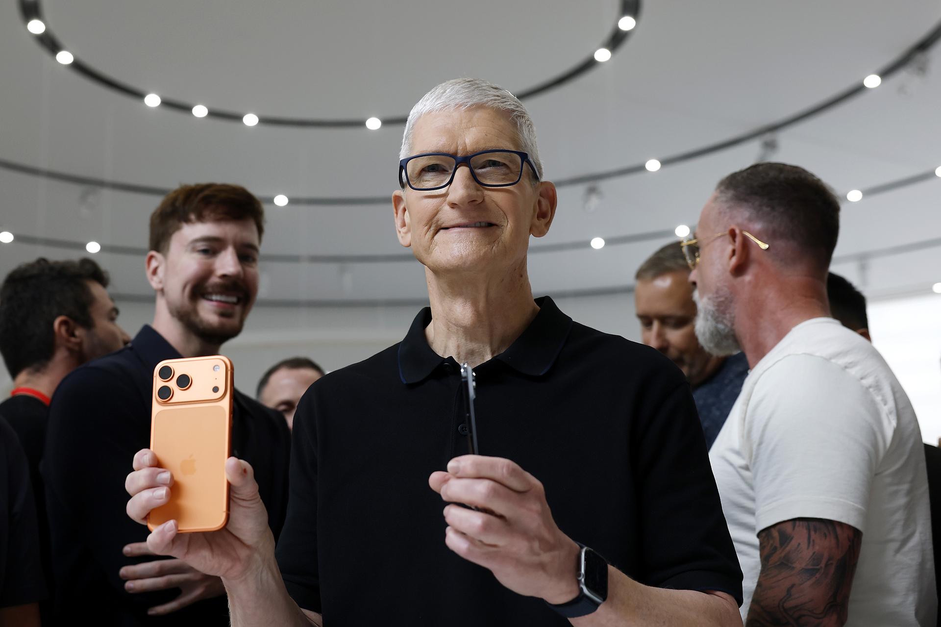 A man with gray hair and black glasses in a black polo shirt holds up a slender copper-colored iPhone.