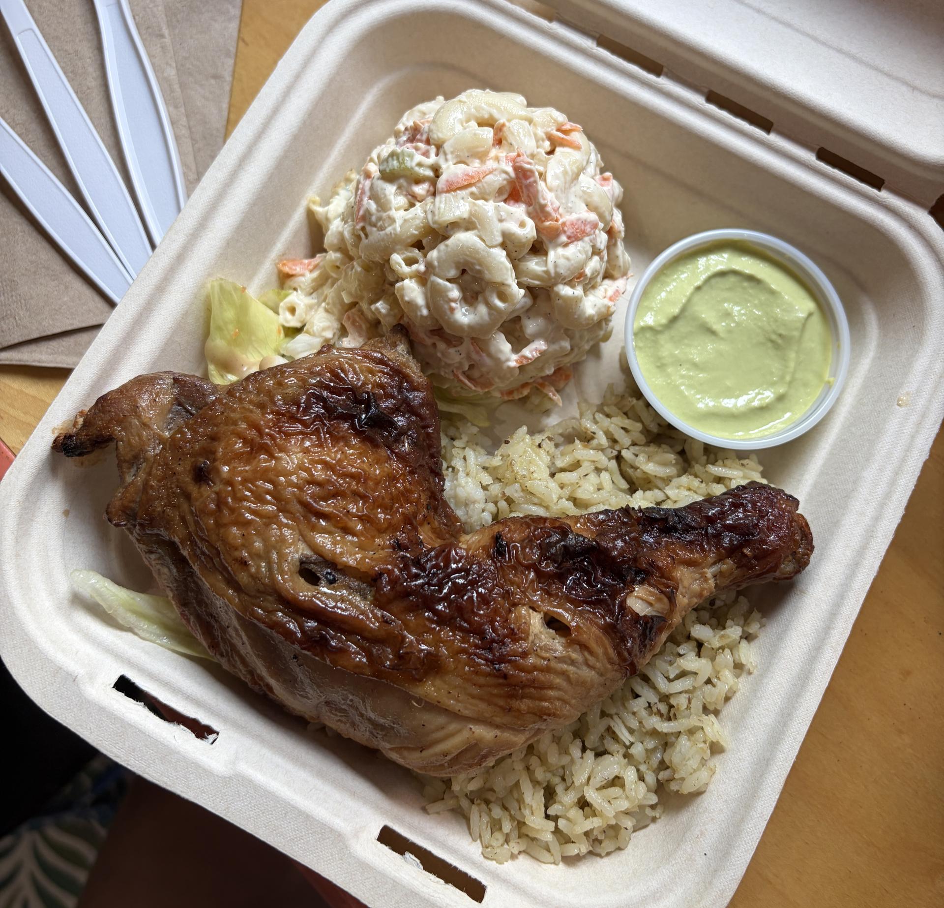A cardboard tray of a big roasted chicken thigh, rice, creamy mac and cheese, and a saucer of green sauce.