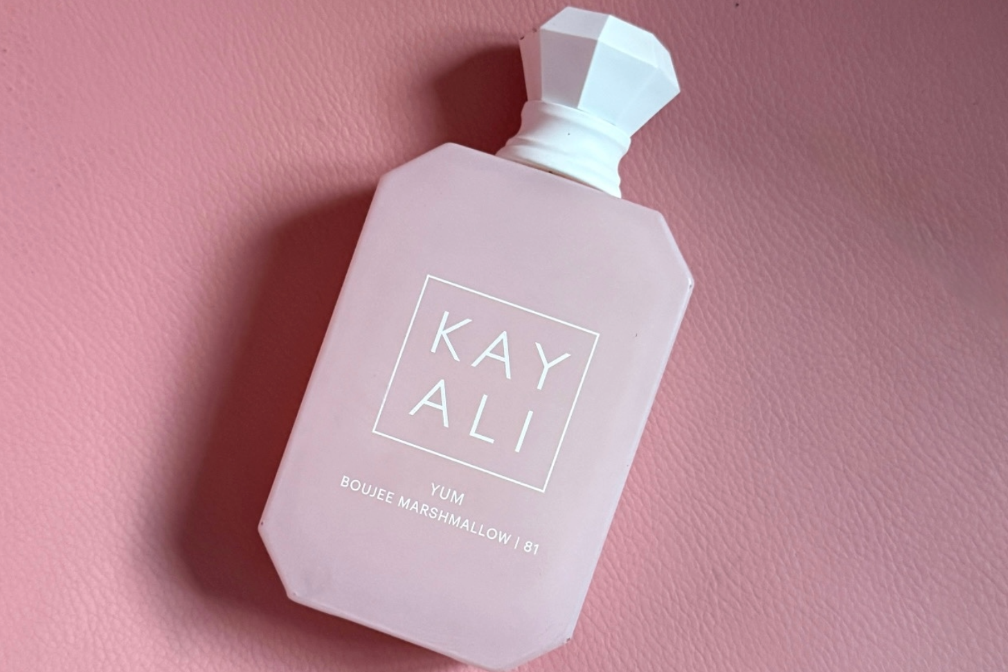 A pink, faceted perfume bottle sits against a pink background, with white text on the front reading 