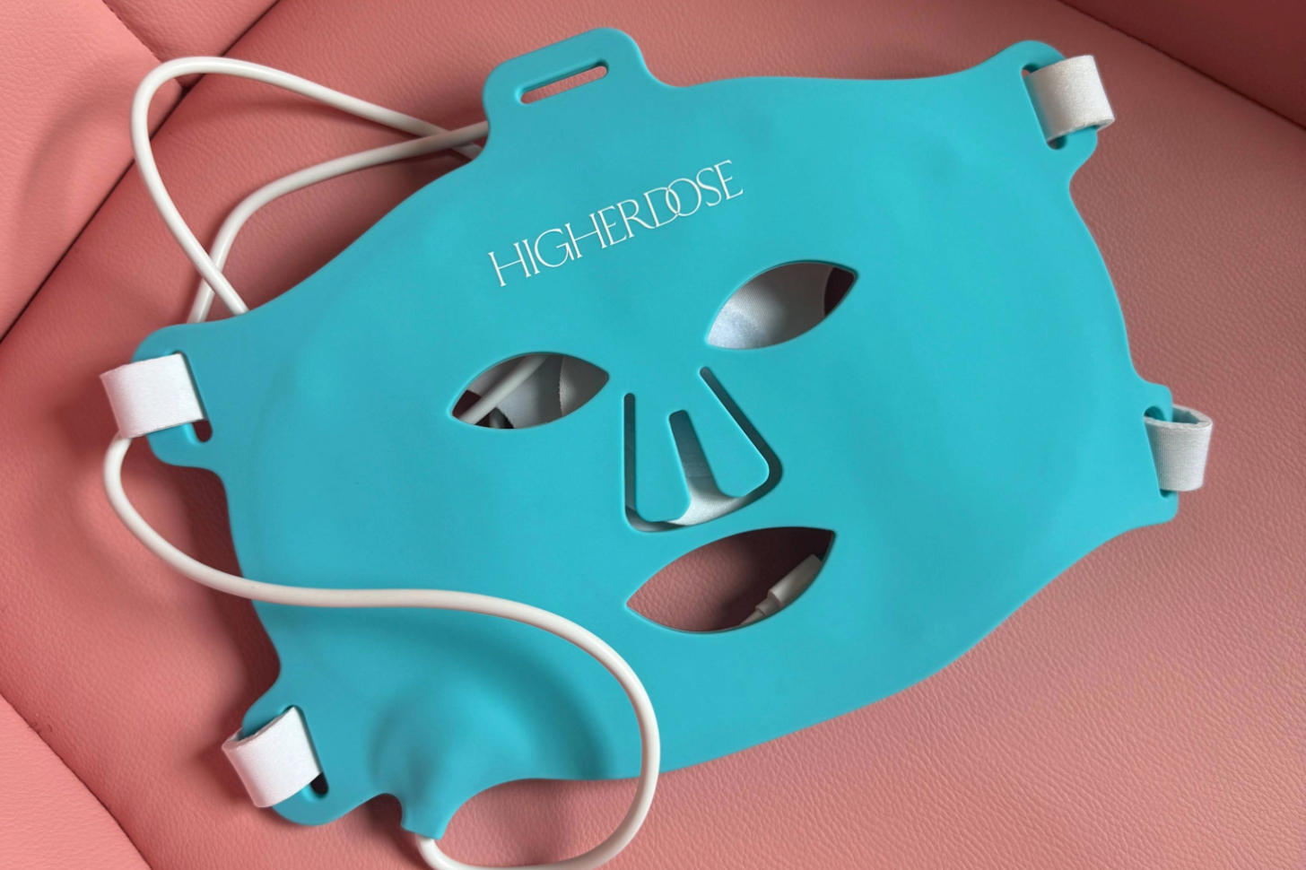 Turquoise HigherDOSE LED face mask with cutouts, resting on a pink surface.