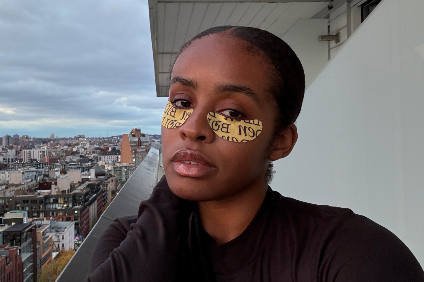 Naomi Parris holds a Miu Miu perfume bottle with a faceted amber base and round black cap against a plain wall.