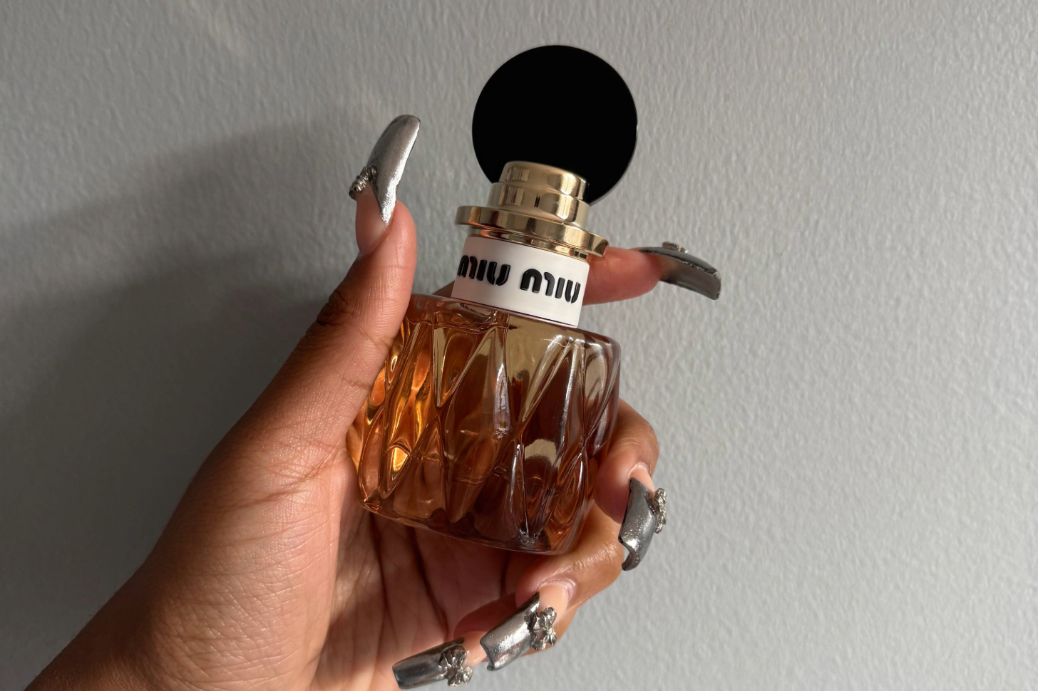 Naomi Parris holds a Miu Miu perfume bottle with a faceted amber base and round black cap against a plain wall.