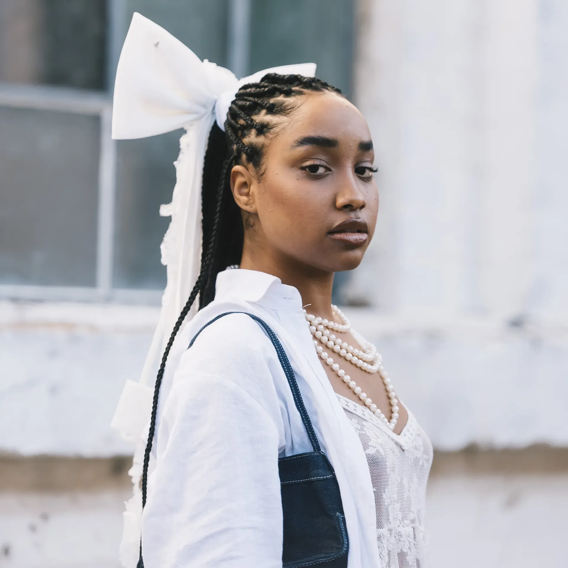 A woman with braided hair styled into a high ponytail tied with an oversized white bow looks toward the camera, wearing a white lace top layered with a sheer white shirt and a pearl necklace. She carries a dark shoulder bag, standing against a softly blurred urban backdrop.
