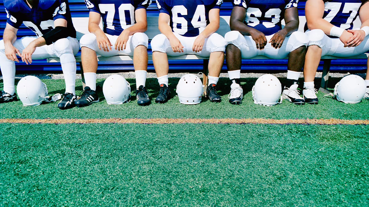 Photo of a football team in uniform sitting on a bench with their helmets in front of them on the field.