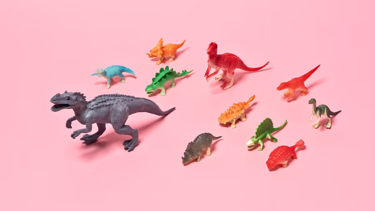 A picture of plastic toy dinosaurs all facing one way on a pink background.