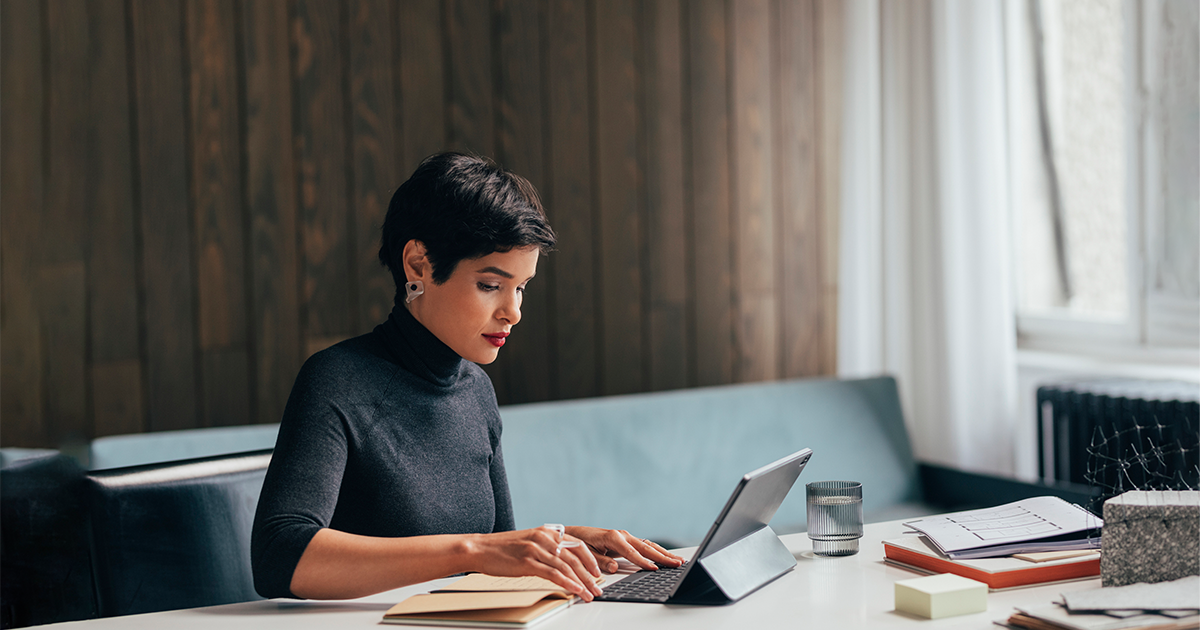 Person seated at a desk, typing on a tablet keyboard with papers, a notebook, and a glass of water nearby.