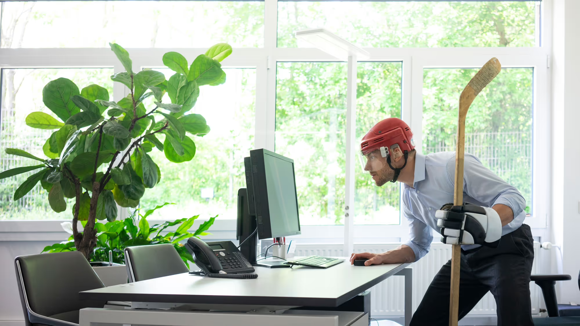 A picture of a person in business attire wearing a helmet and holding a hockey stick while looking at their computer.