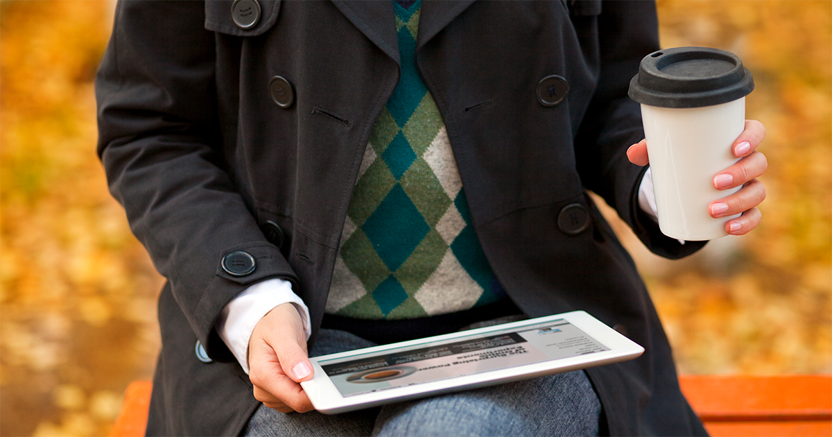 Person in black coat holds a tablet and takeaway coffee cup while seated on an orange bench. Autumn leaves scattered on the ground suggest a cozy fall setting.
