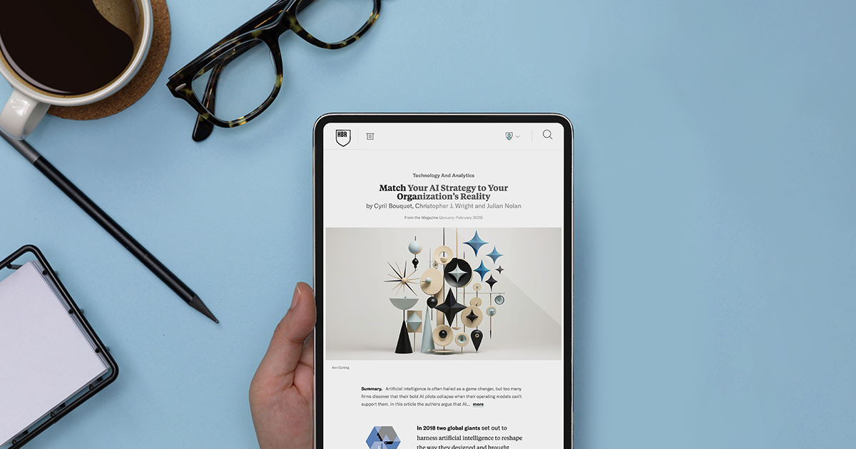 Tablet displaying an HBR article about AI strategy on a blue desktop with coffee, glasses, and a notebook.