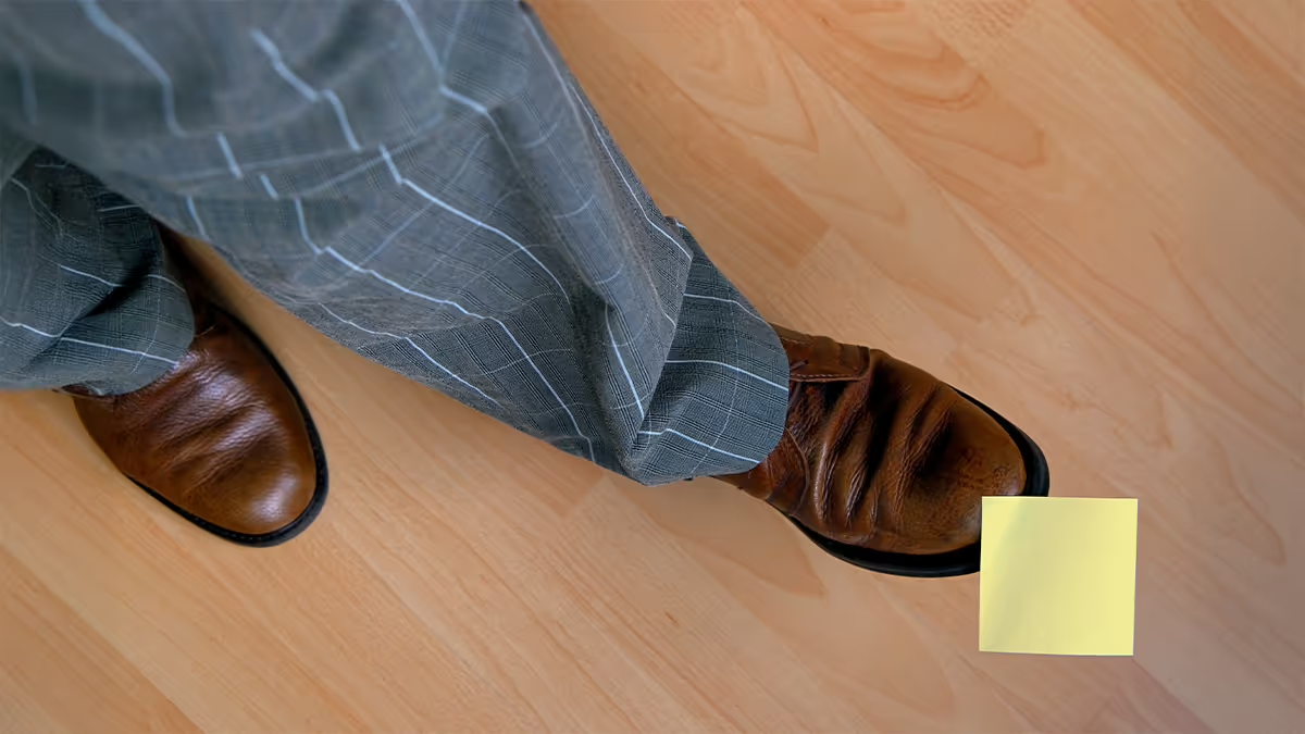 Person wearing gray plaid pants and brown leather shoes standing on a light wooden floor, with a yellow sticky note placed near the right shoe.