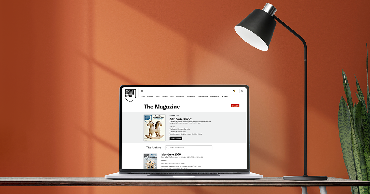 Laptop displaying HBR’s Magazine page on a wooden desk, beside a black lamp and tall green plant against a warm orange wall.