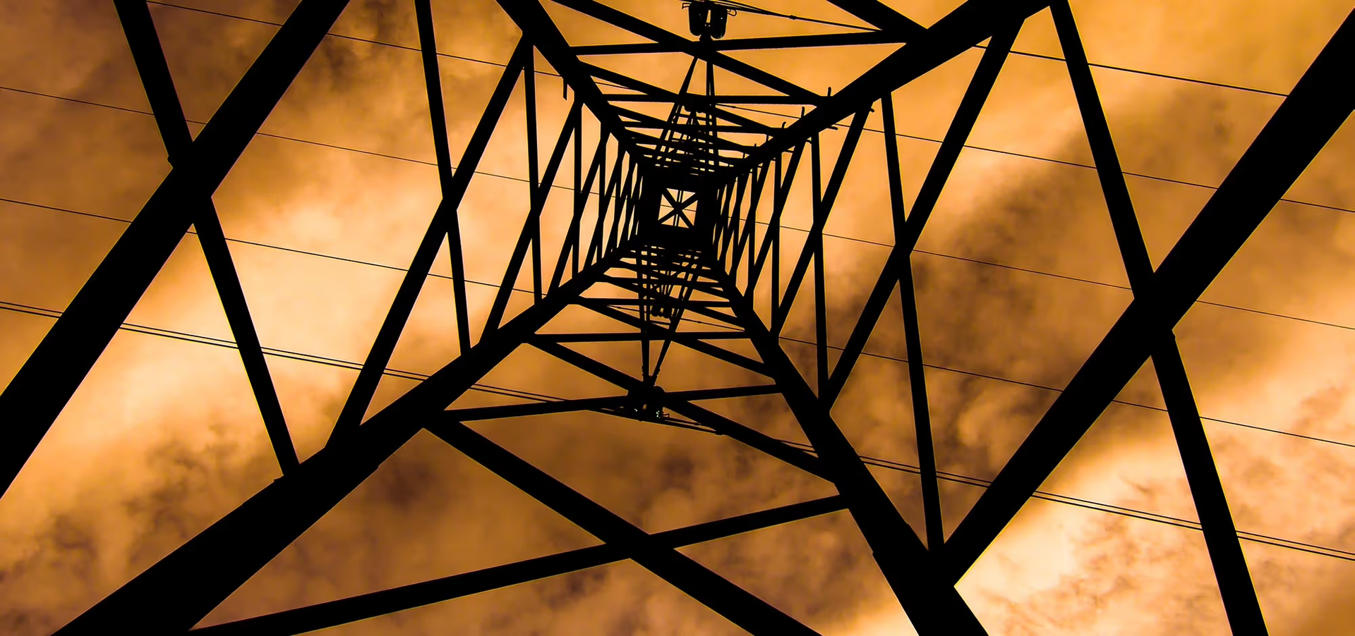 View looking up inside a metal transmission tower, with geometric beams silhouetted against an orange sky.
