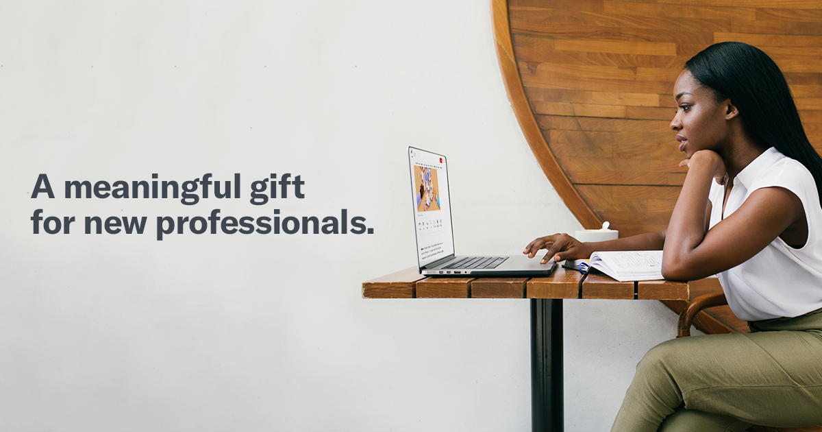 A woman sits at a cafe table, focused on her laptop, with a notebook beside her. Text on the wall reads, 