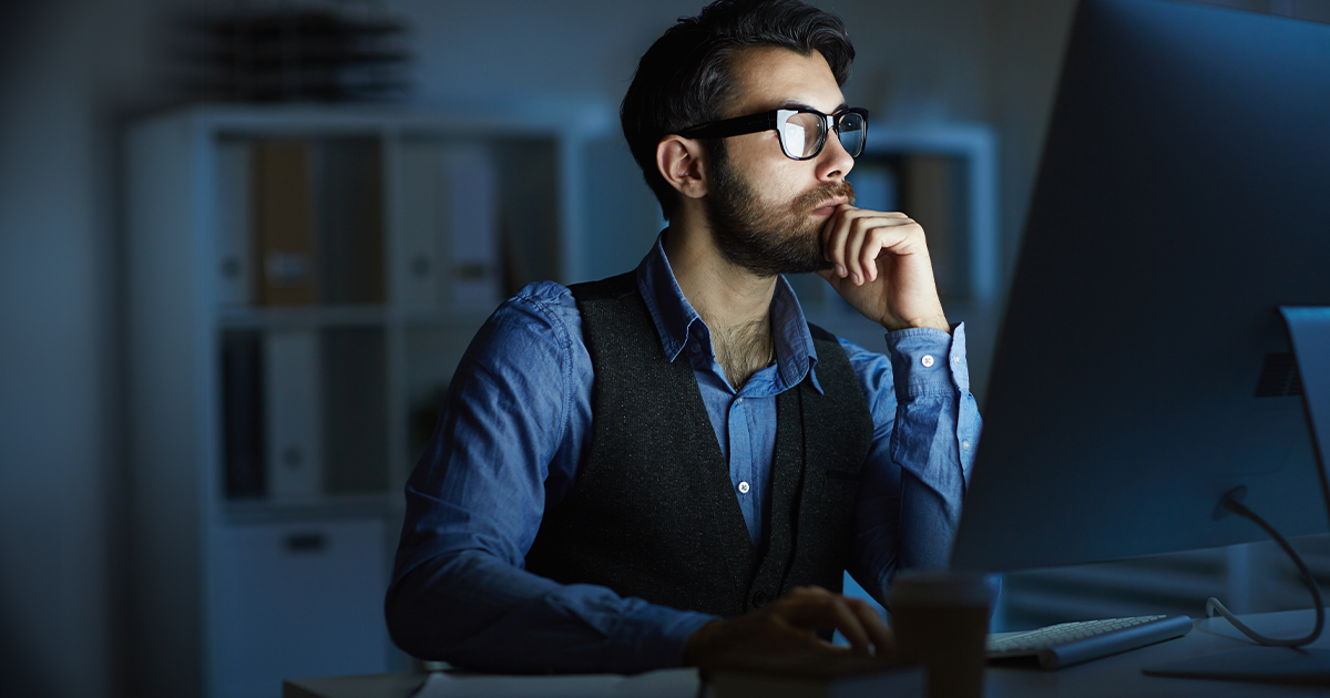 Person wearing glasses sits at a computer workstation, looking at a large monitor in a low-lit office setting.