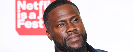 Kevin Hart wears a blue jacket and looks off to the side while photographed on a red carpet.