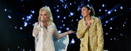 Dolly Parton in a white gown singing alongside Miley Cyrus in a gold oversized suit at the 61st Grammy Awards. 