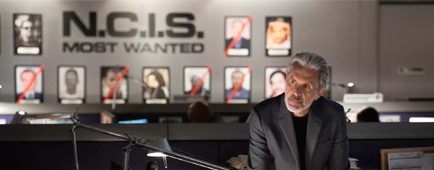 A man standing at a desk with a wall with the wording "NCIS most wanted" and rows of photos seen behind him.