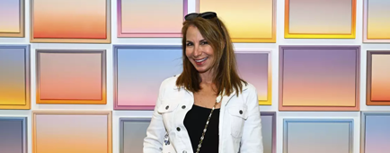 Jill Zarin in a white jacket, black top and jeans posing in front of a tiled backdrop.