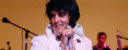 A man with dark hair, in a white jacket and high collars, holds a mic while smiling and looking to the left A man with dark hair, in a white jacket and high collars, holds a mic while smiling and looking to the left