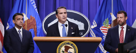 A man with dark hair, in a dark suit and blue tie, speaks at a lectern, flanked by two other men, also in suits and ties